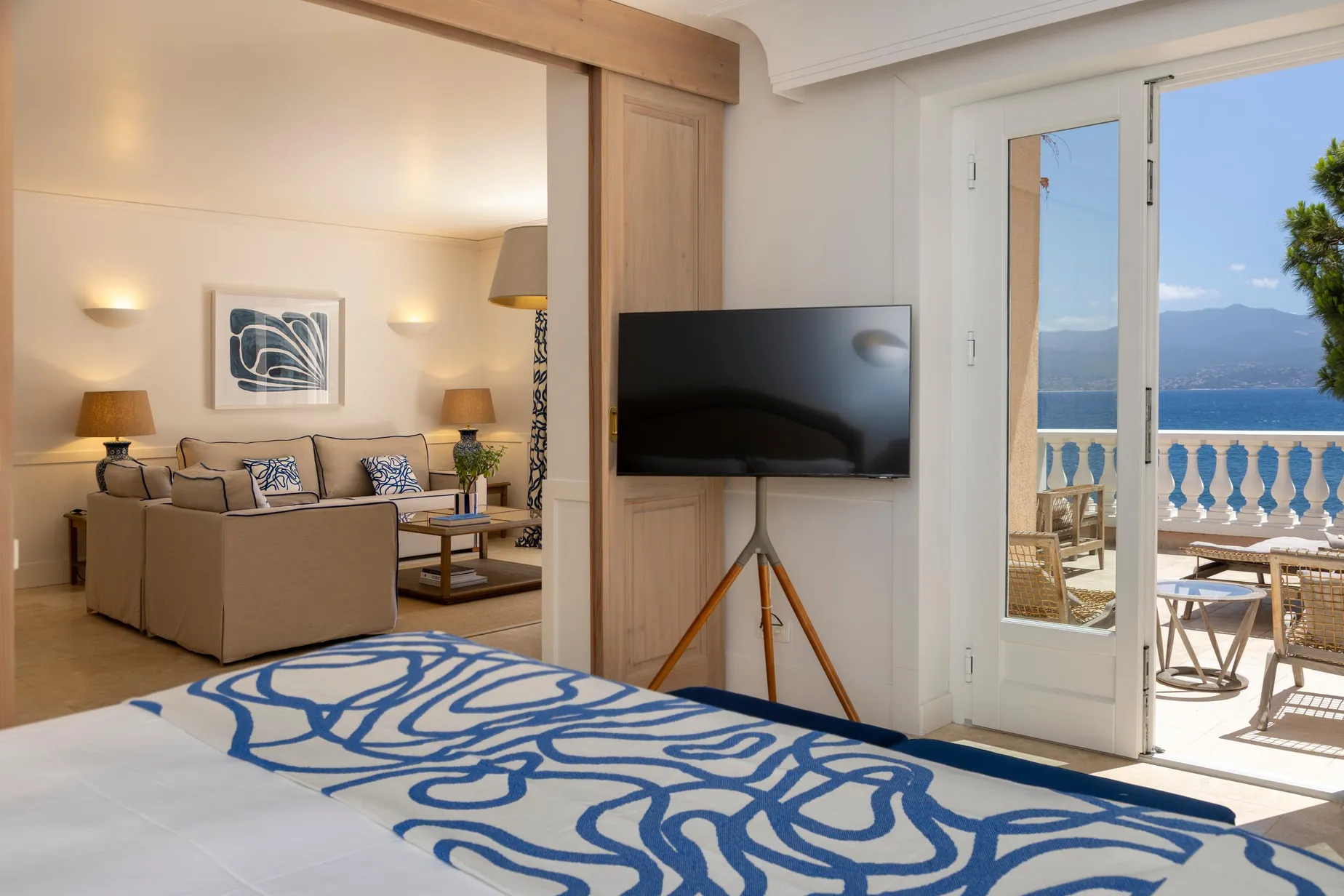Zimmer mit blau-weiß gemustertem Bett, Sofa, Fernseher auf Stativ und Blick auf Balkon mit Meerblick in der Ferne