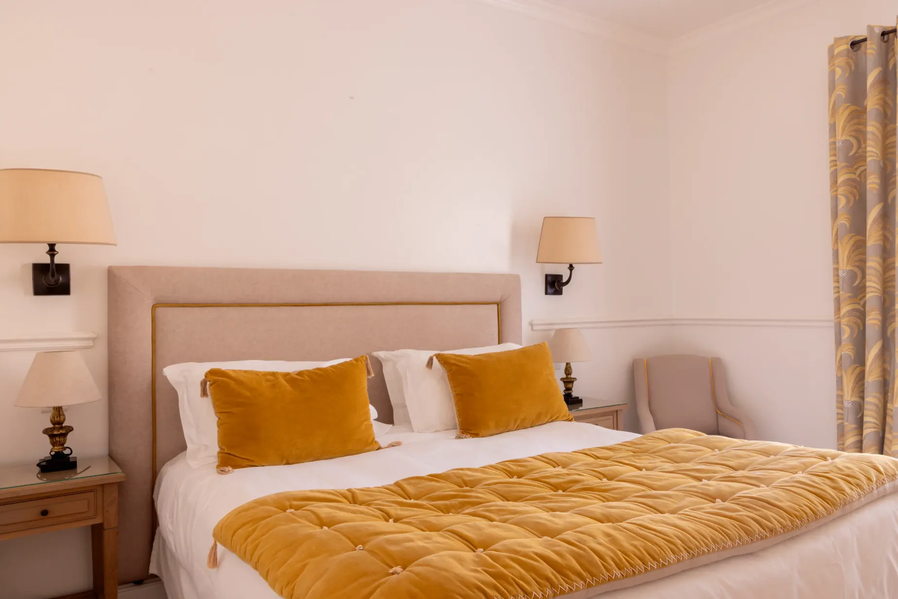 Lit double avec une tête de lit beige, une couverture et des coussins dorés, deux tables de chevet avec lampes et des rideaux à motifs