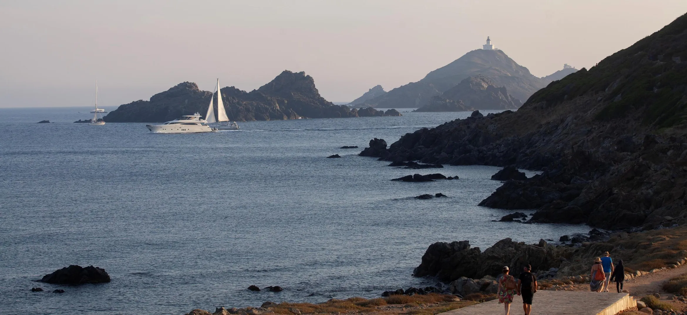 Un sentier côtier avec des promeneurs longe une mer calme bordée de rochers, avec des voiliers et un phare en arrière-plan