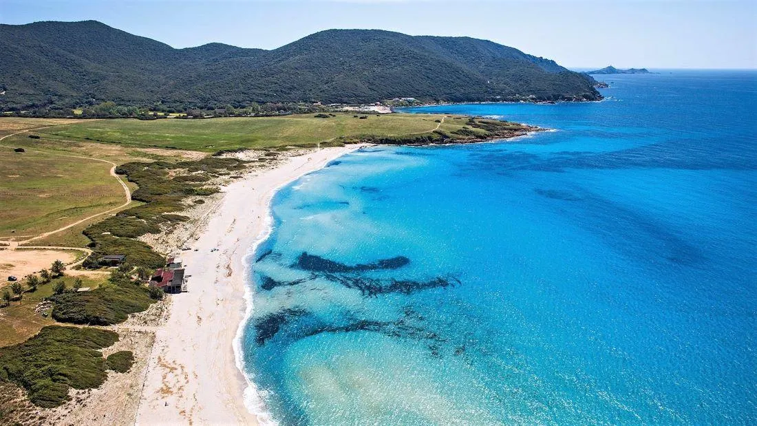 Vue aérienne d'une plage de sable blanc bordée d'une mer turquoise, avec des collines verdoyantes en arrière-plan