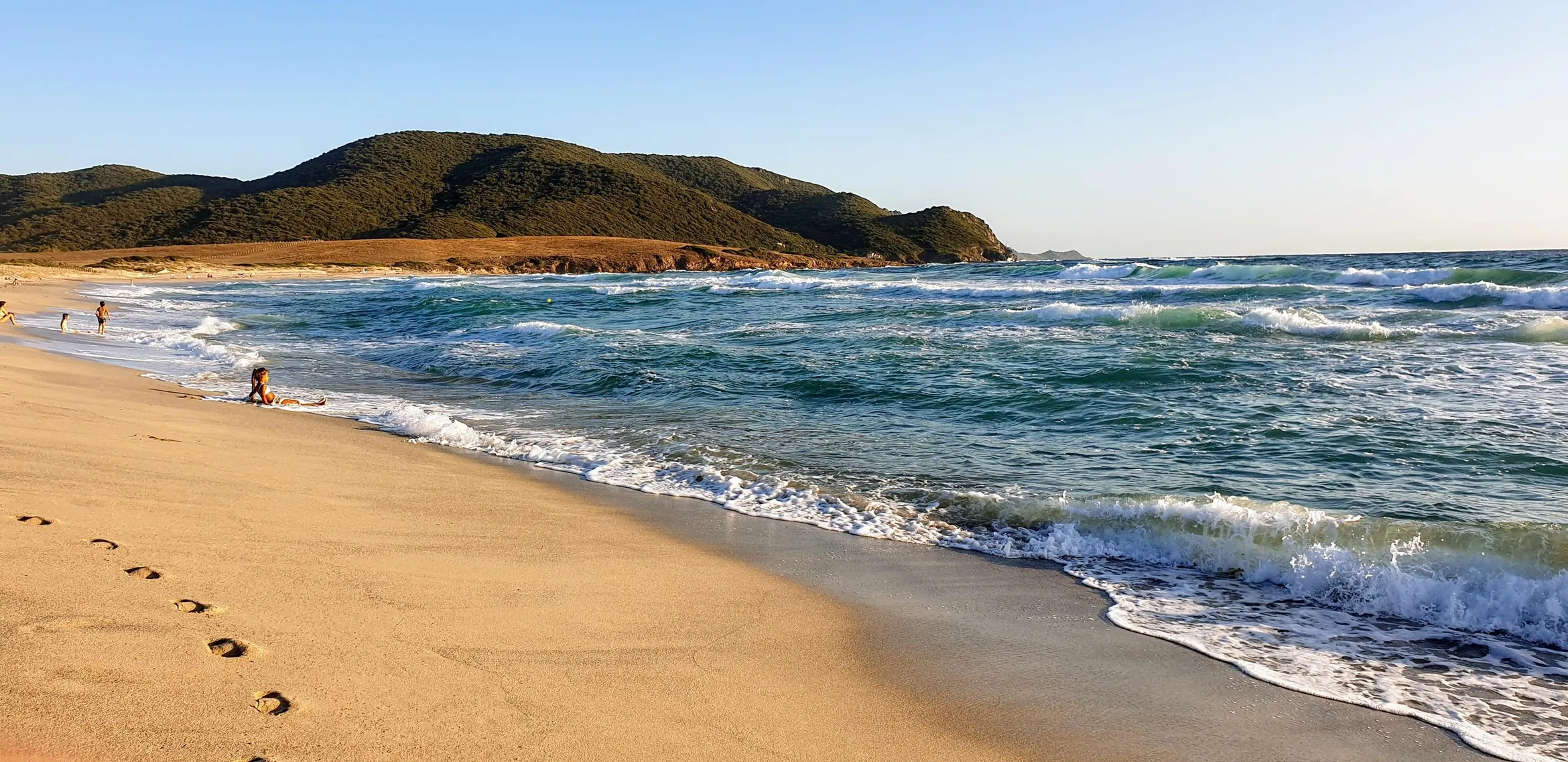 Sandstrand mit einigen Personen, Ozeanwellen und grünen Hügeln im Hintergrund
