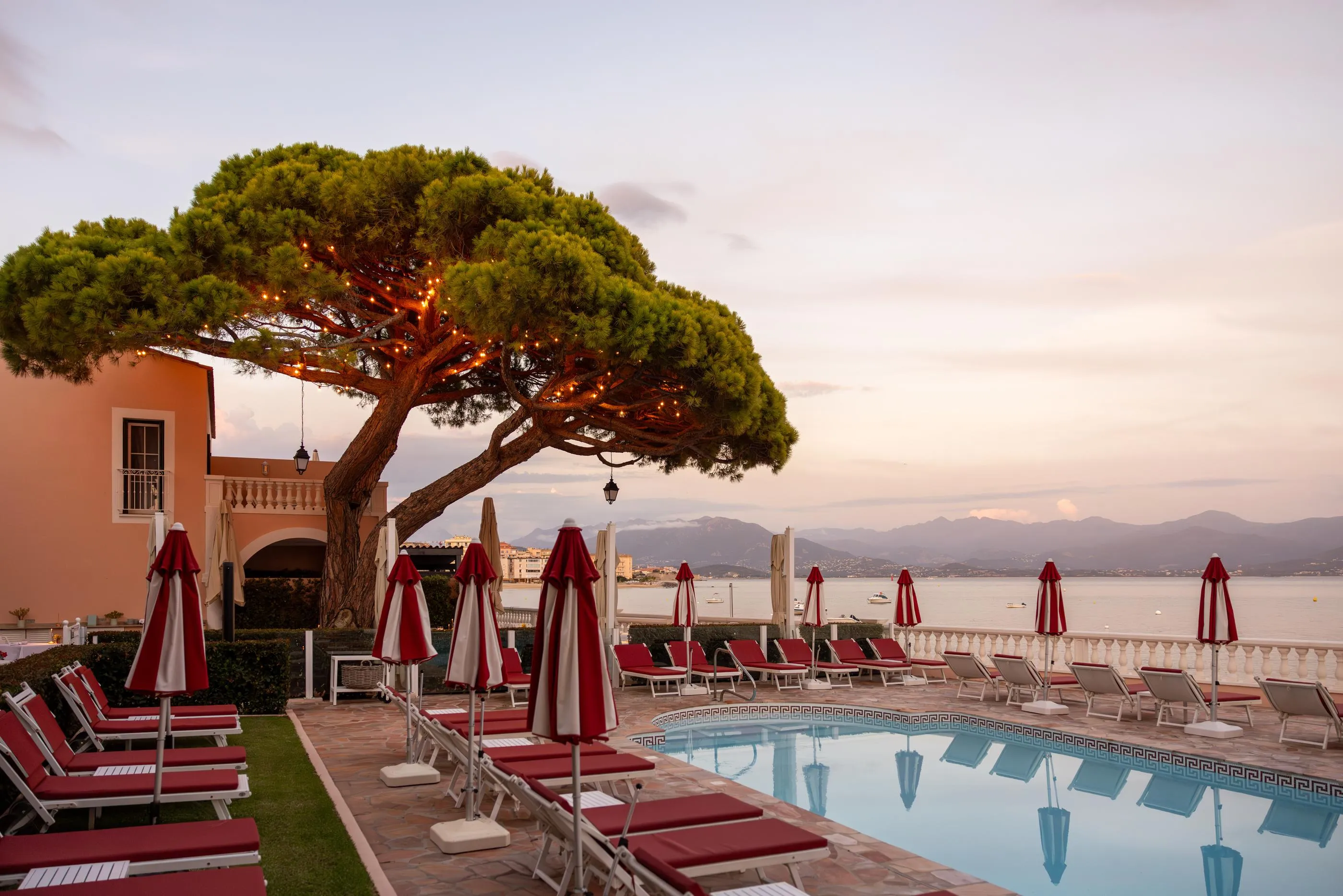 Piscine avec transats et parasols rouges, située sur une terrasse face à la mer, avec un arbre illuminé et un bâtiment en arrière-plan