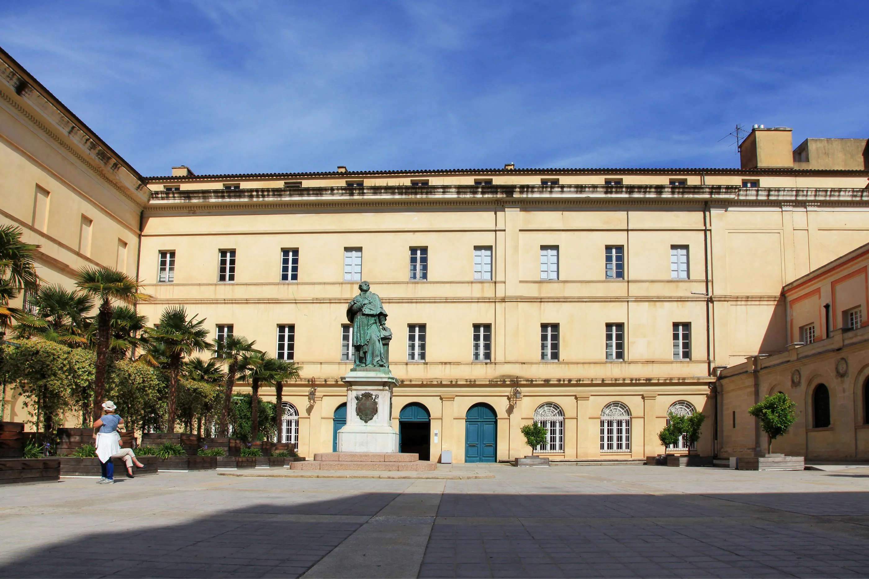 Courtyard with a two-story beige building, windows, and a central statue on a pedestal