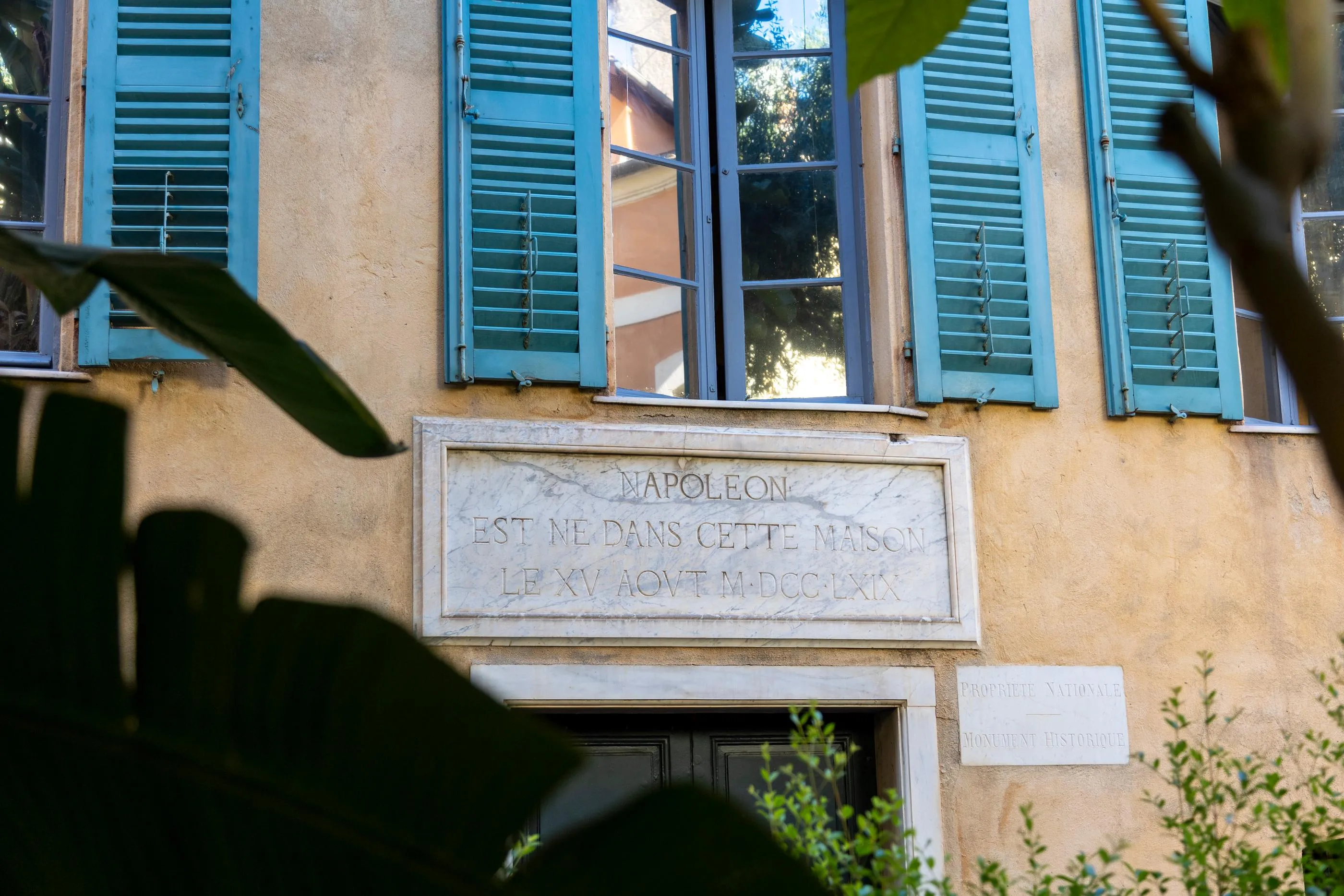 Commemorative plaque on a yellow wall indicating "Napoleon was born in this house on August 15, 1769" with closed blue shutters in the background