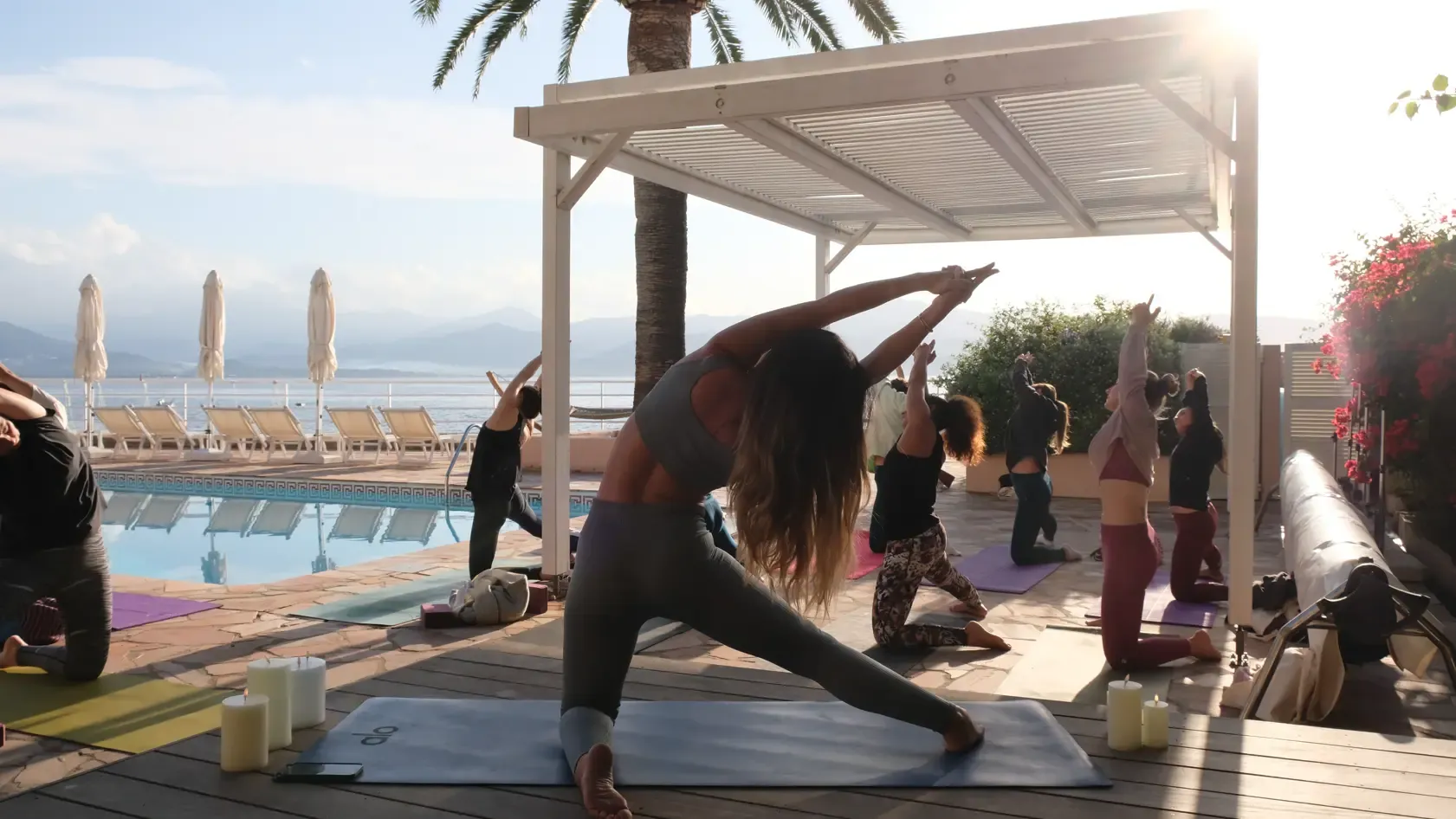 Un groupe de personnes pratique le yoga en extérieur près d'une piscine avec une vue sur la mer et des montagnes au loin