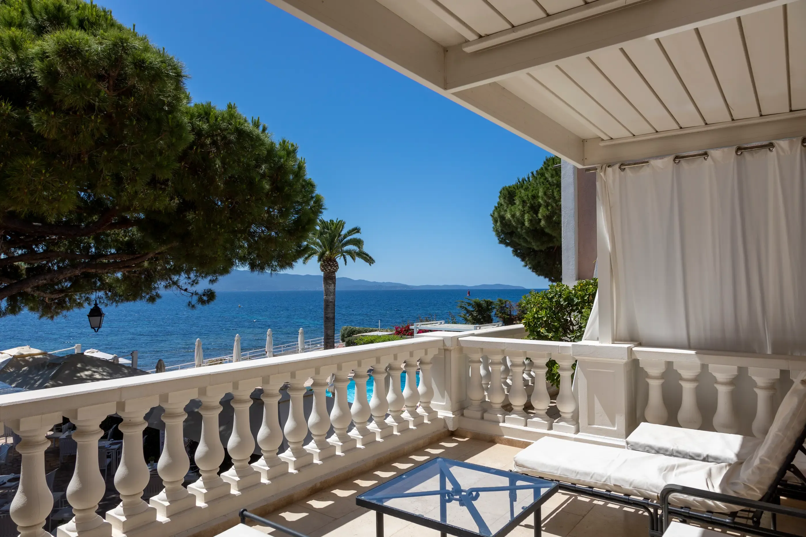 Balcon avec vue sur la mer, équipé de chaises longues et d'une table, entouré d'une balustrade en pierre et de rideaux blancs