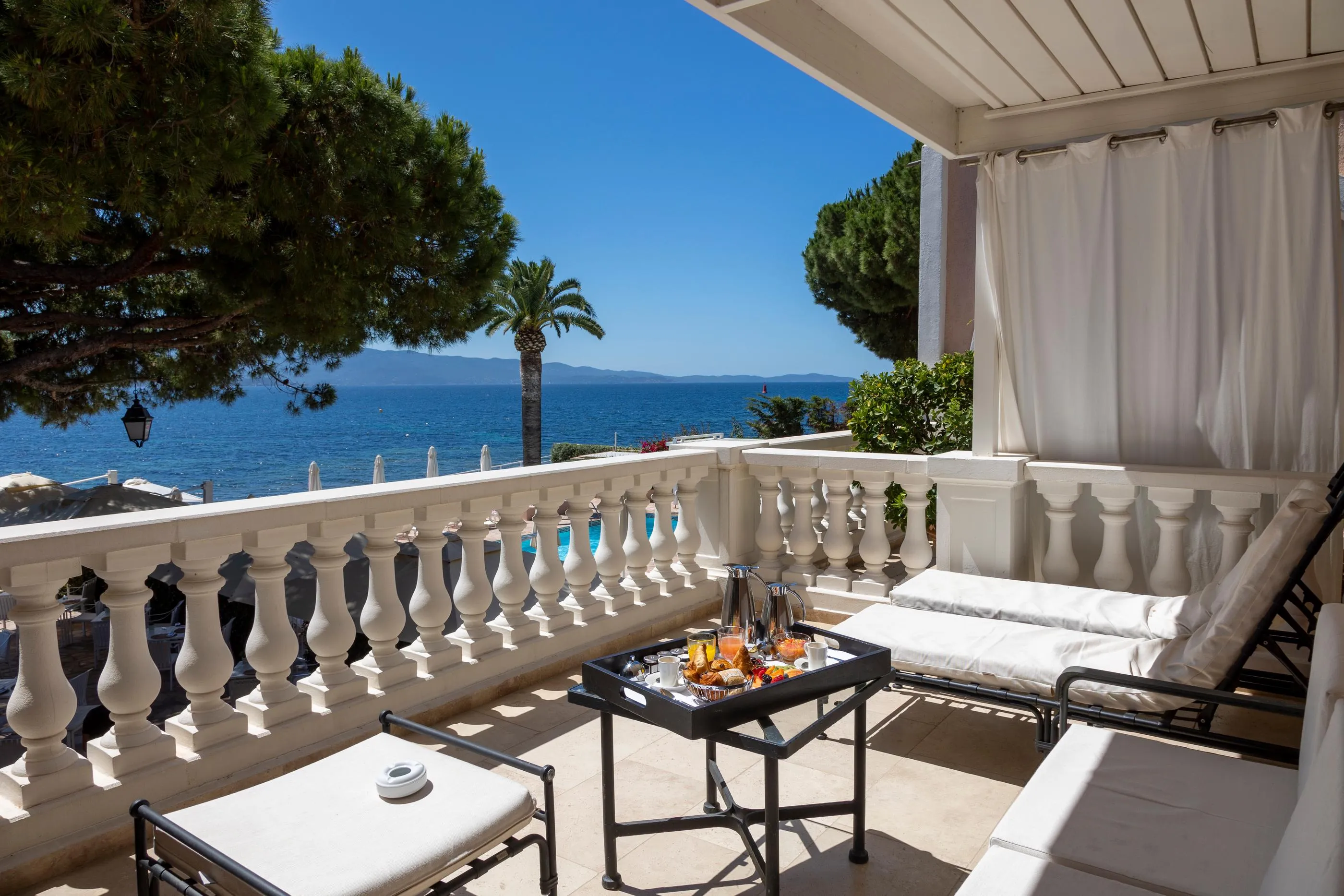 Balkon mit Meerblick, ausgestattet mit Liegestühlen, einem Tisch mit Getränken und Essen, umgeben von einer Steinbalustrade und weißen Vorhängen
