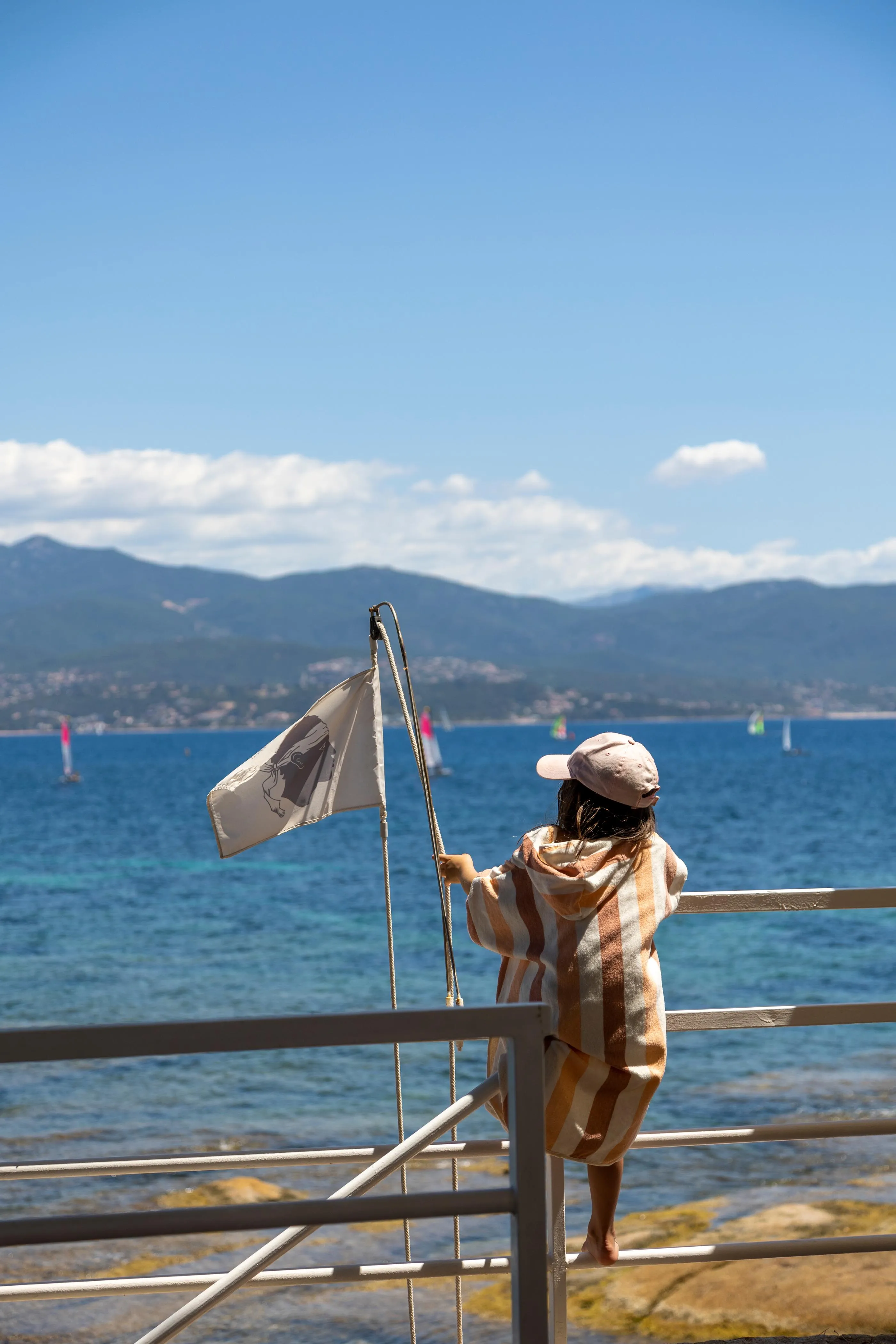 Une personne en robe et chapeau tient une bannière sur un balcon face à un lac et des montagnes