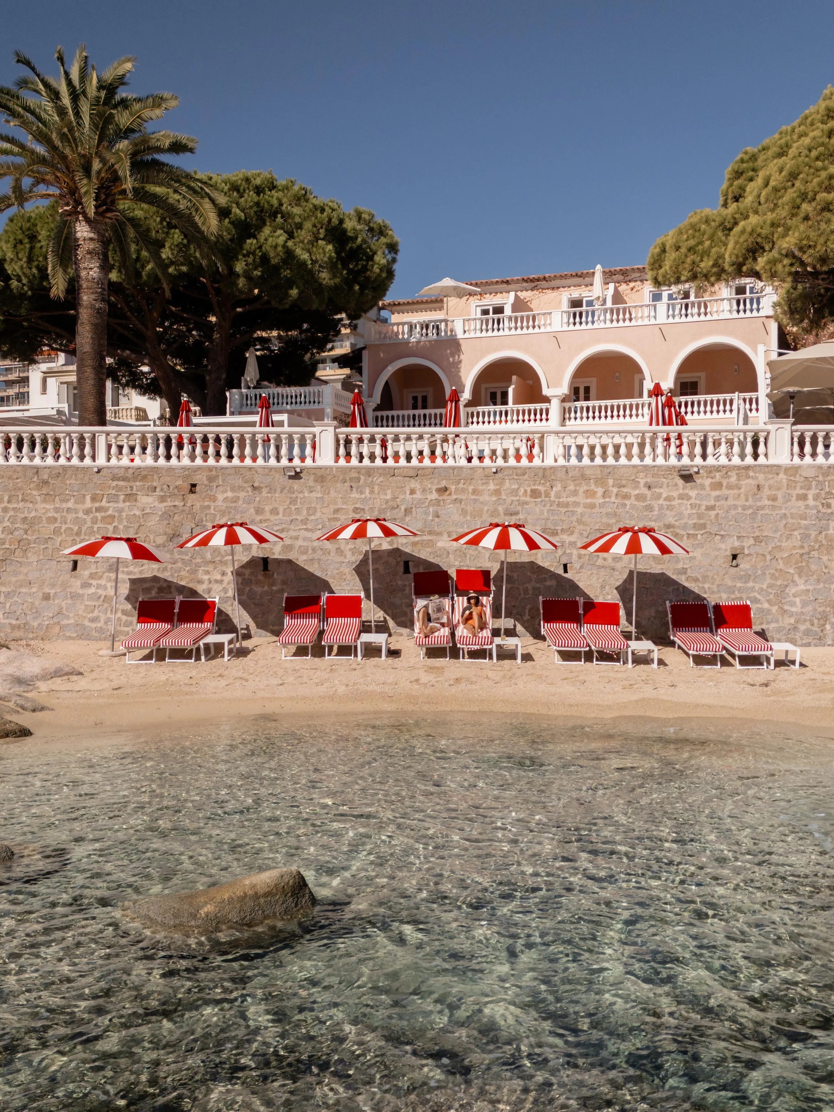 Une plage avec des transats et parasols rayés rouges et blancs devant un bâtiment à arcades et balustrade blanche