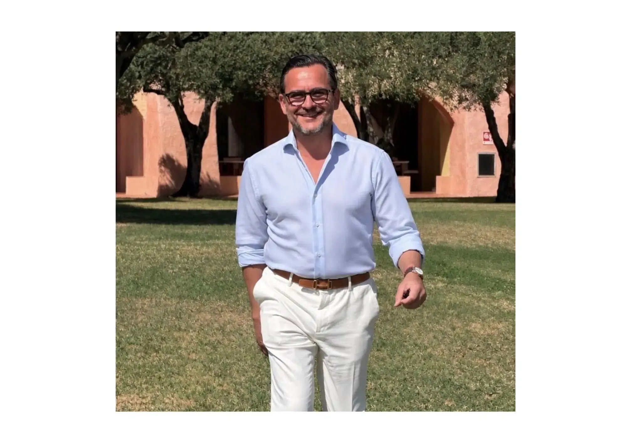 Smiling man in blue shirt and white pants, standing on a lawn in front of a pink building