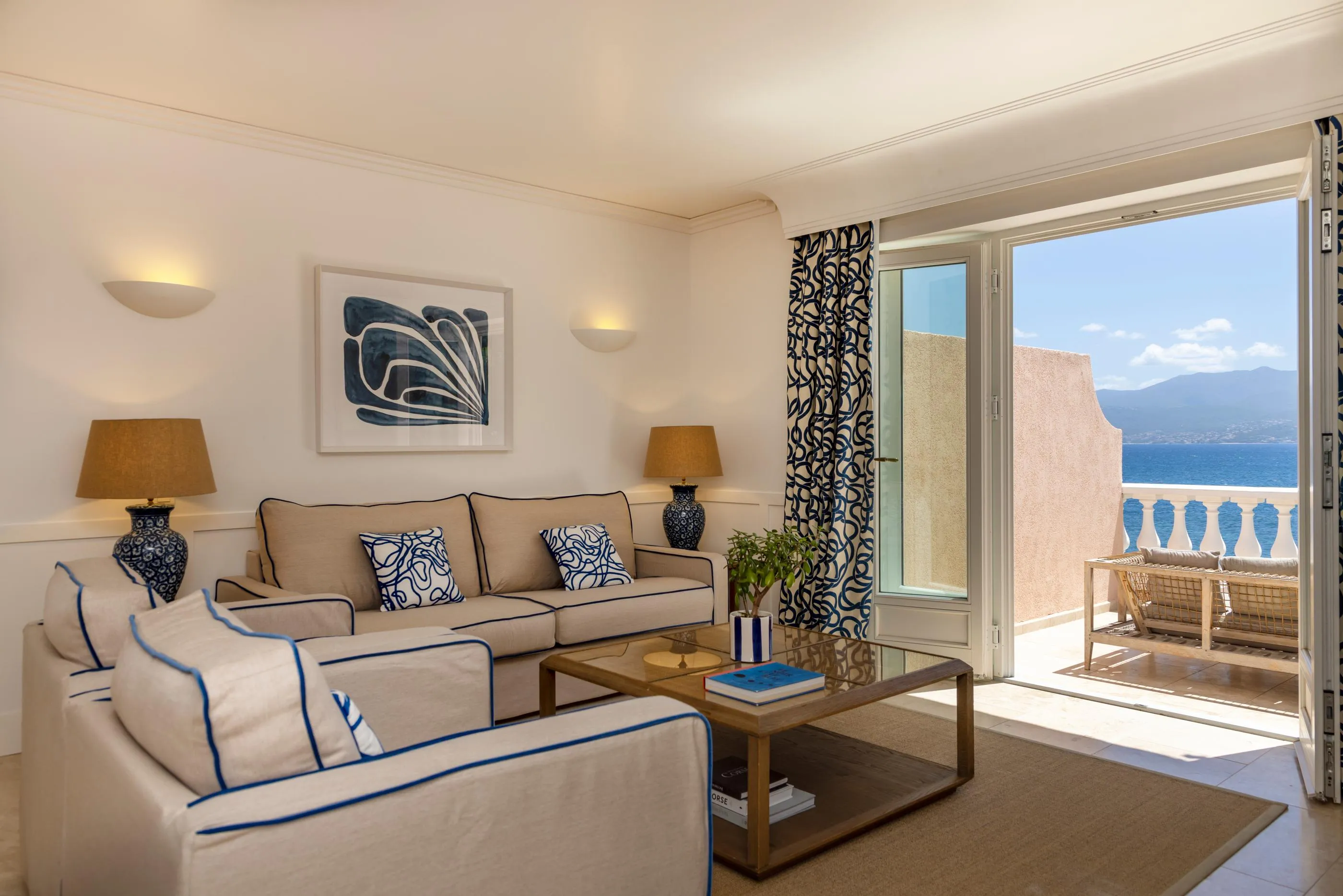 Wohnzimmer mit beigem Sofa mit blauen Kanten, Glastisch, Meerblick durch eine offene Terrassentür