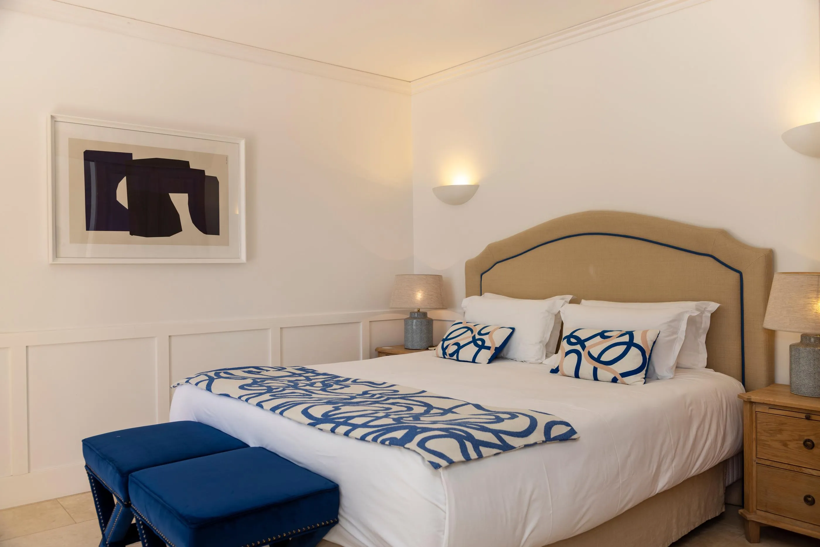 Zimmer mit einem Doppelbett, das mit einer weißen Decke und einer blauen, gemusterten Decke bedeckt ist, zwei Nachttischen mit Lampen, einem schwarzen und weißen abstrakten Bild an der Wand und einer blauen Bank am Fußende des Bettes.