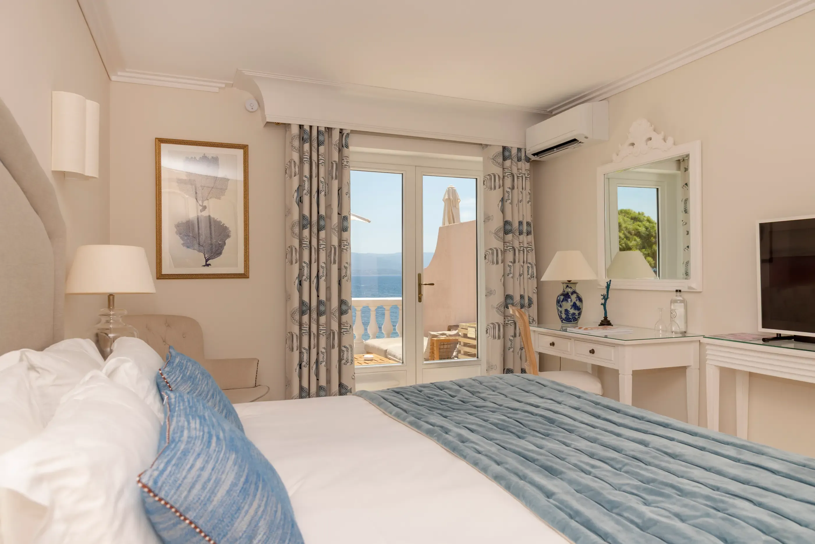 Zimmer mit einem Bett, das mit einer blauen Decke bedeckt ist, einer Balkontür mit Meerblick, einem Spiegel, einem Fernseher und einer Nachttischlampe.