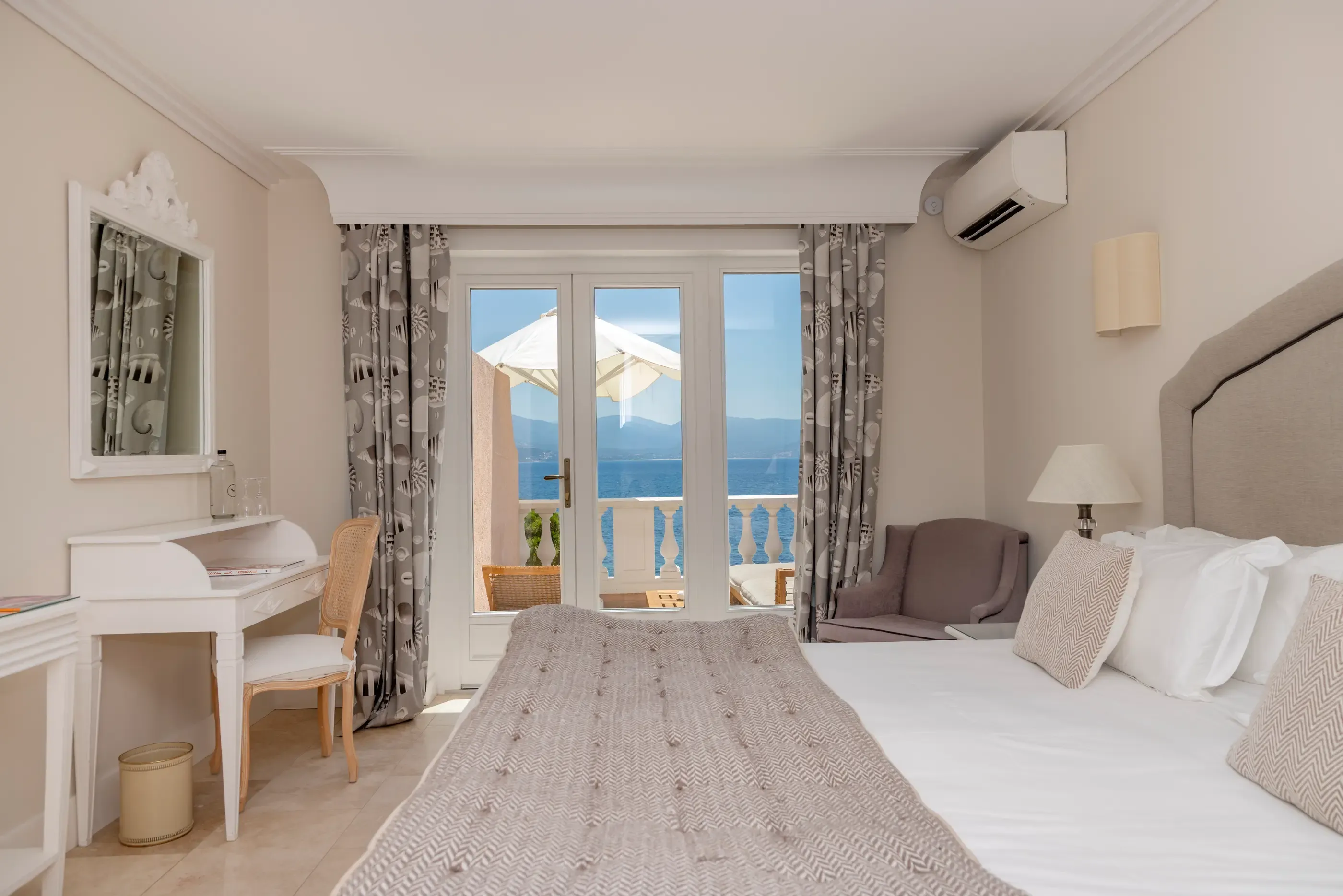 Zimmer mit Doppelbett, Schreibtisch, Balkontür mit Zugang zu einem Balkon mit Meerblick und Bergblick, graue Vorhänge und sanfte Beleuchtung