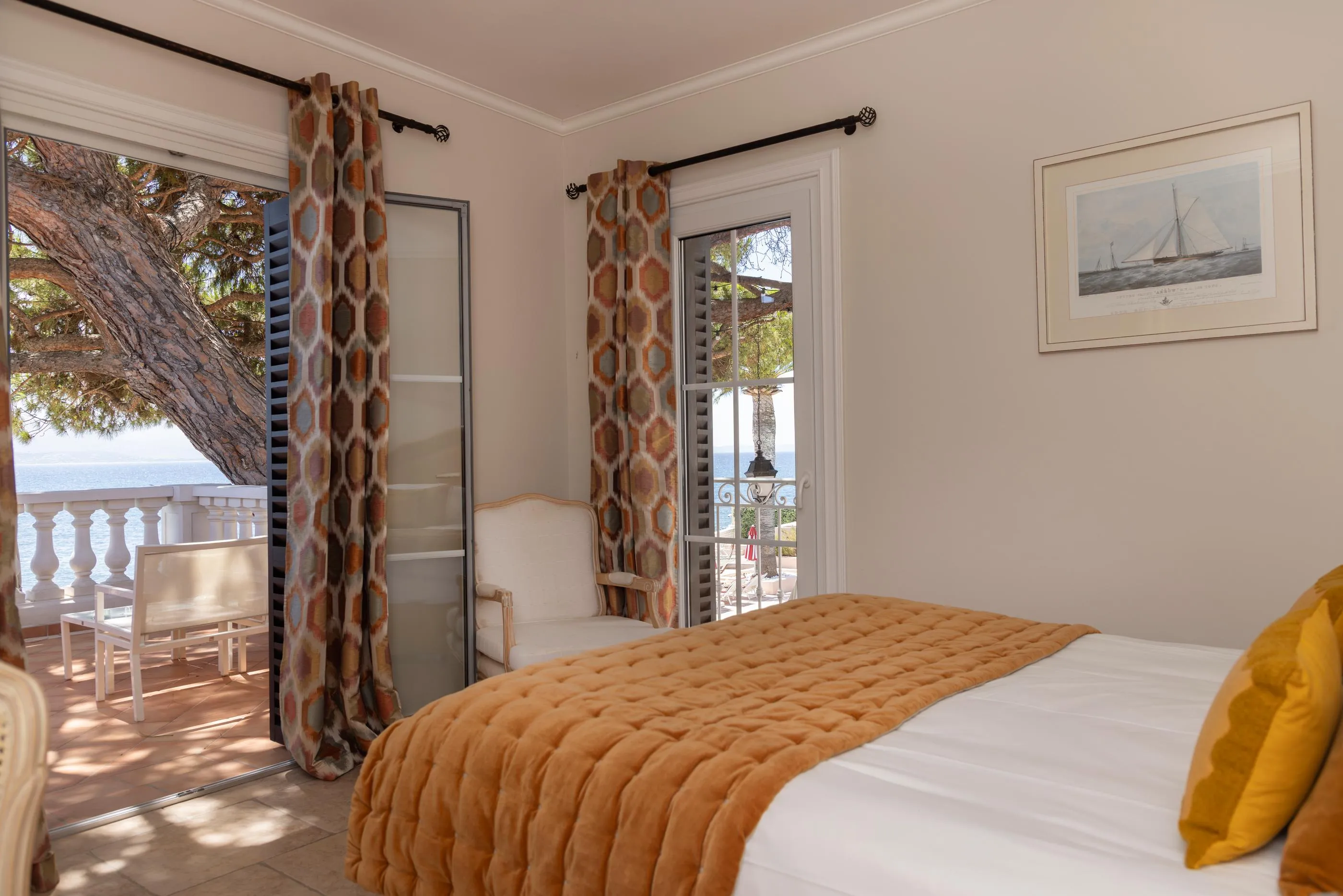 Zimmer mit einem Bett, das mit einer orangen Decke und einem gelben Kissen bedeckt ist, Blick auf einen Balkon mit Stühlen und das Meer in der Ferne, ein Bild eines Segelboots an der Wand