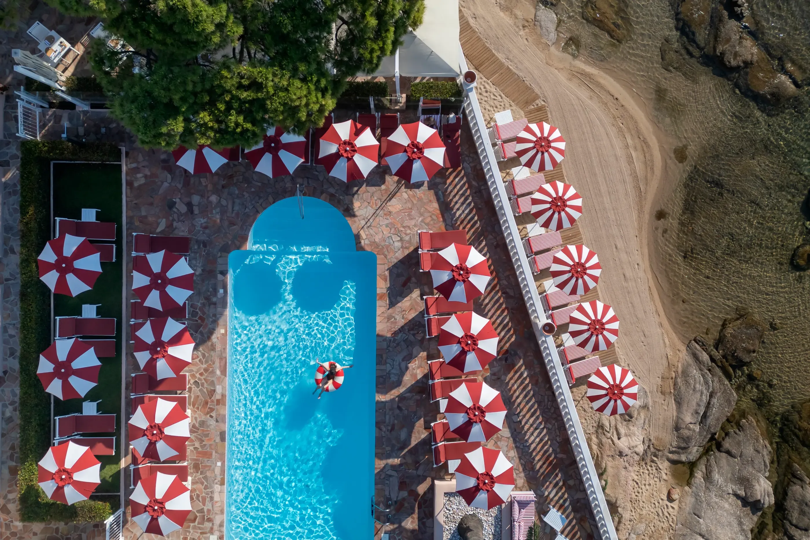 Vue aérienne d'une piscine entourée de transats et de parasols rouges et blancs, située près d'une falaise et d'une zone boisée